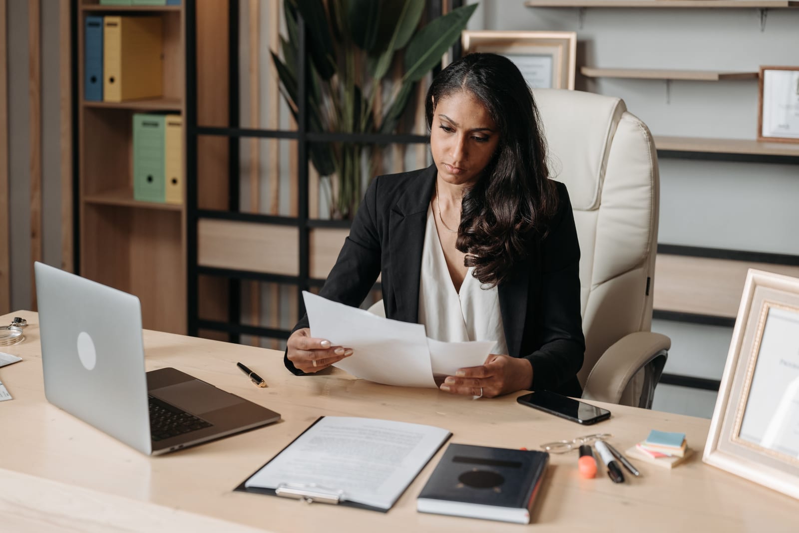 A woman at an office desk reads a sheet of paper, with a laptop, a clipboard of documents, and a shelf of file binders around her