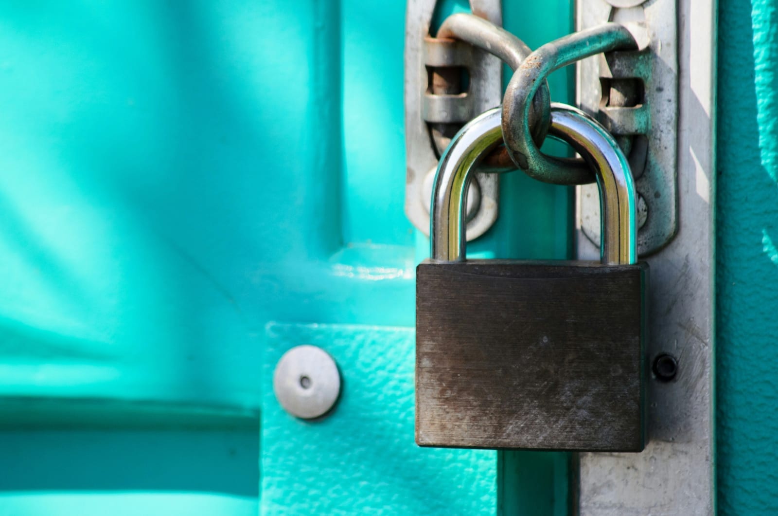 A closed dark metal padlock hanging from a steel hasp on a bright turquoise painted door