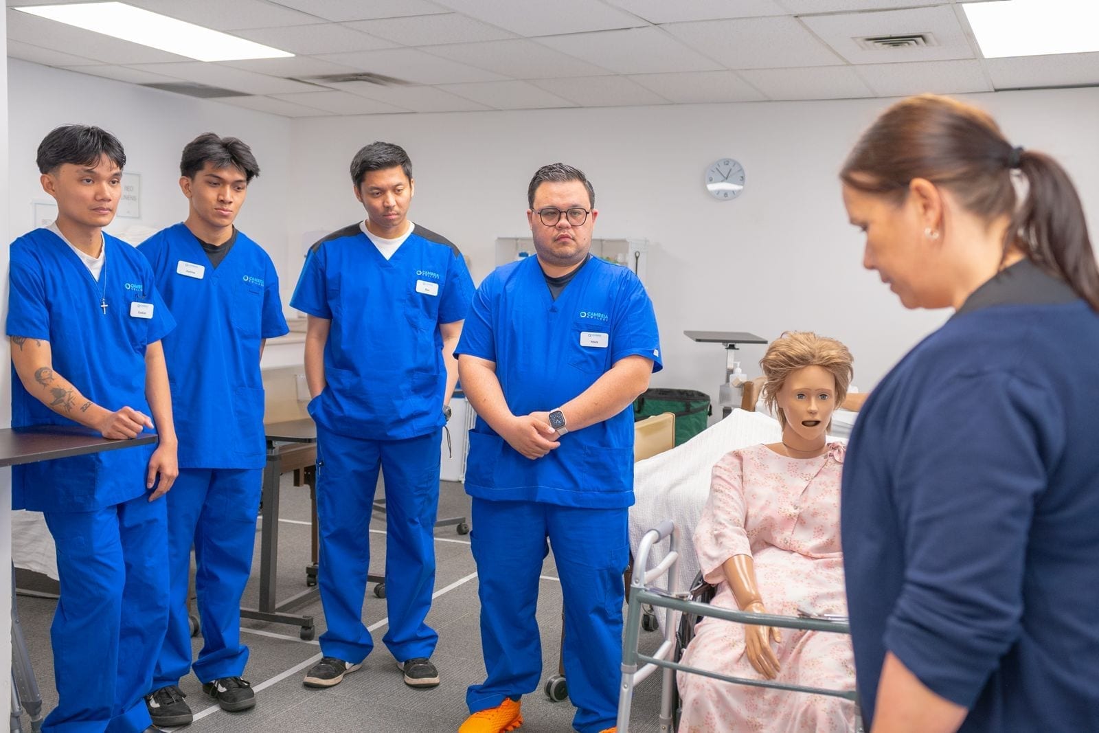 Nursing students in blue scrubs practicing patient care technique with a training mannequin
